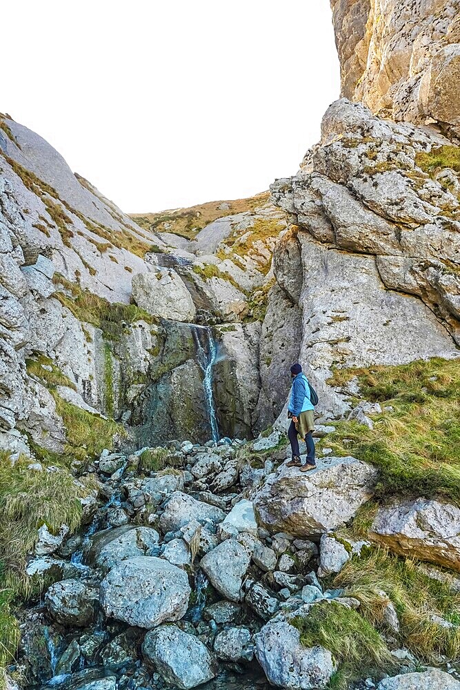 Hiker in blue jacket navigating rocky landscape near a small waterfall on mount txindoki, showcasing the rugged beauty of gipuzkoa, basque country