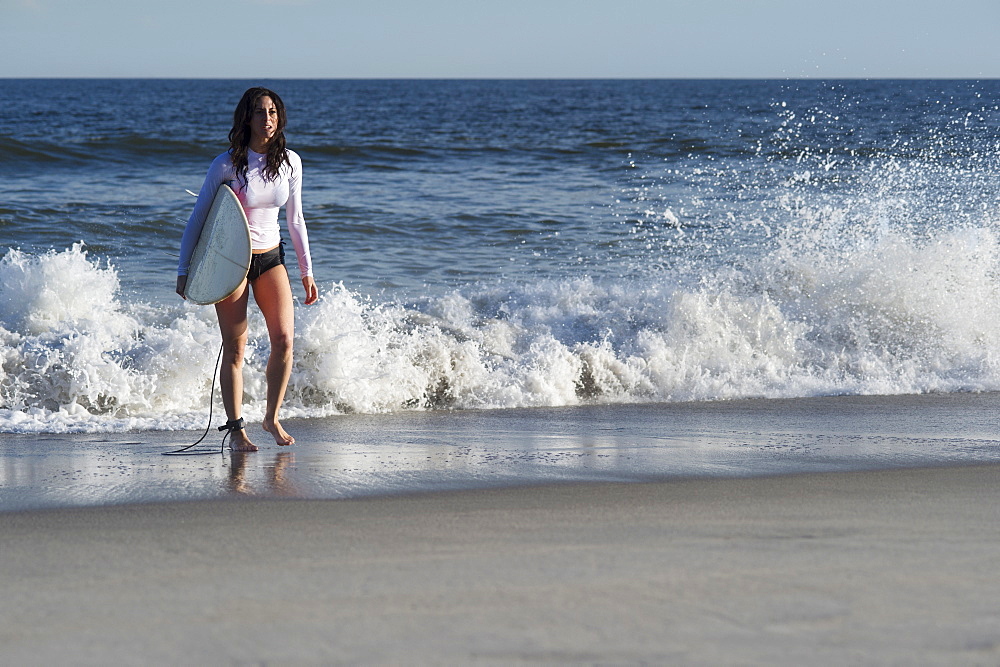 Woman carrying surfboard on beach, Rockaway Beach, New York