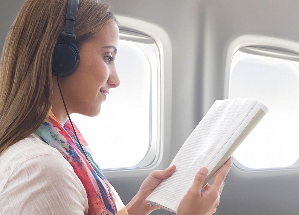 Young woman wearing headphones while reading book on plane