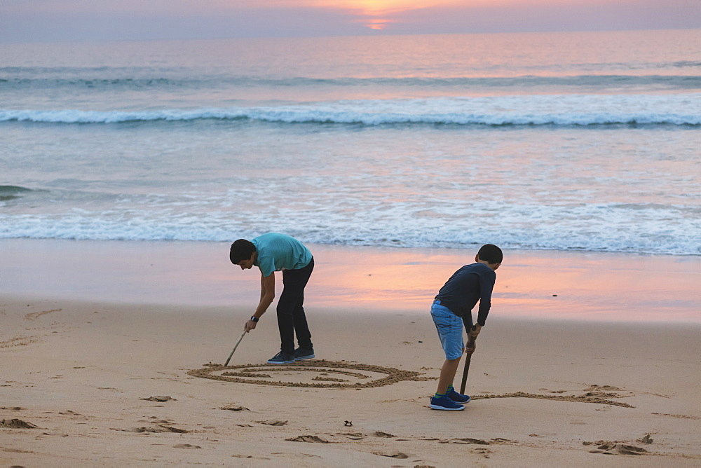 Brothers drawing in sand at sunset