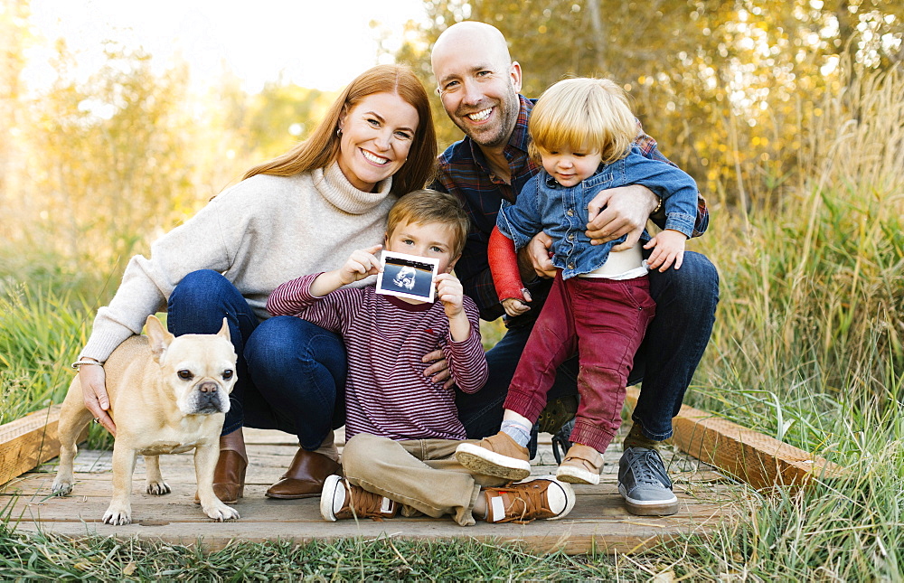 Smiling family with ultrasound photograph and pet dog on forest boardwalk