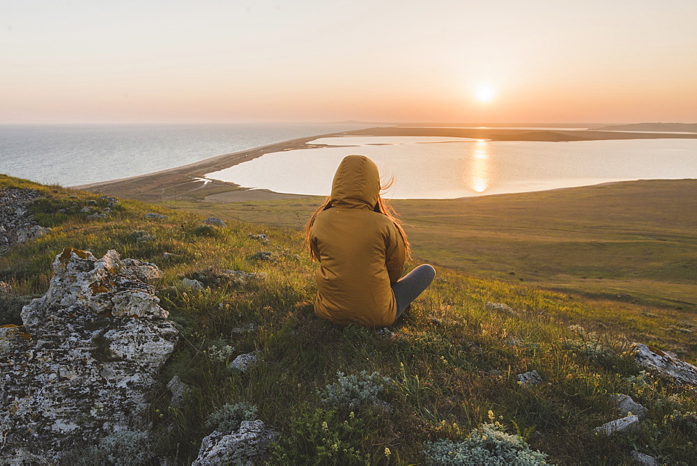 Woman in hooded jacket sitting on hill during sunset