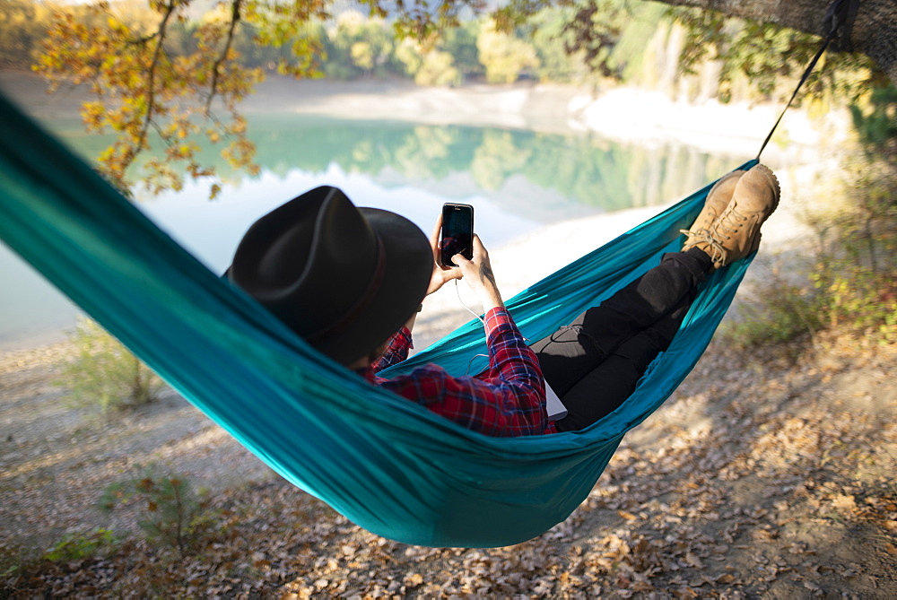 Italy, Man lying in hammock near lake and using smart phone