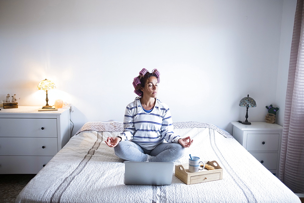 Woman with hair curlers meditating on bed in front of laptop