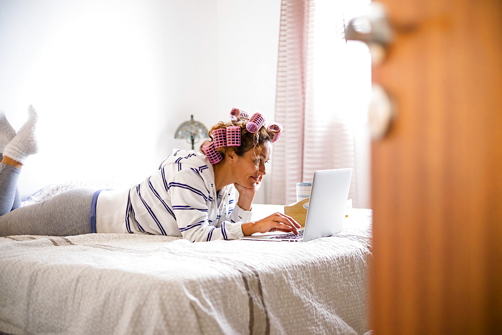 Woman with hair curlers lying on bed and using laptop