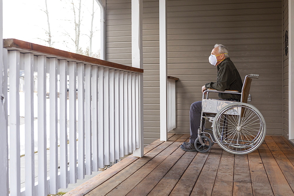 Senior man in wheelchair wearing protective mask to prevent coronavirus transmission on porch