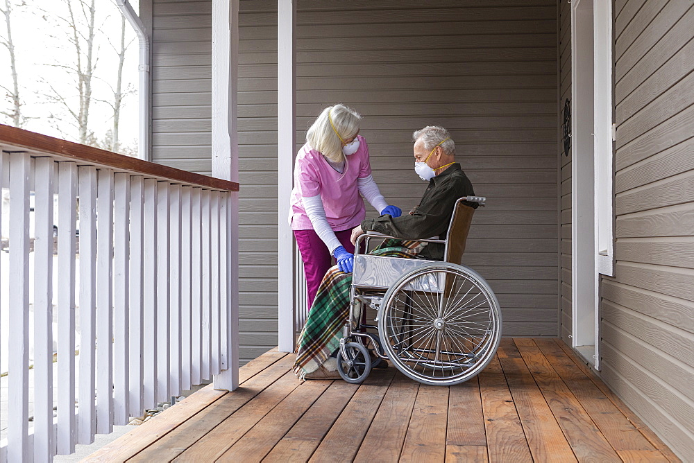 Woman and man in wheelchair wearing protective mask to prevent coronavirus transmission on porch