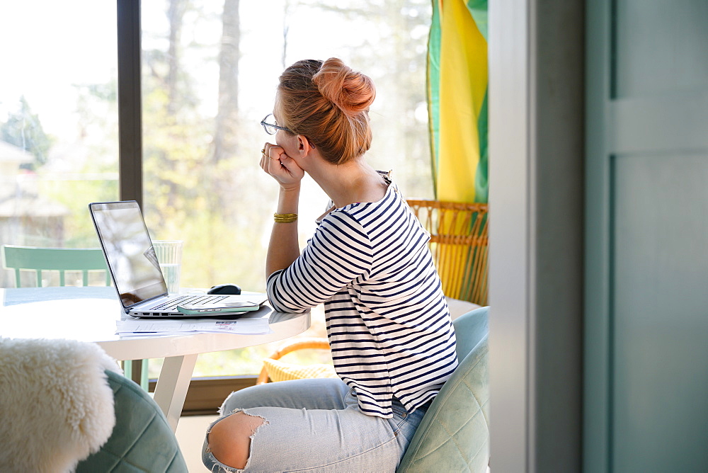 Woman sitting in front of laptop, looking through window