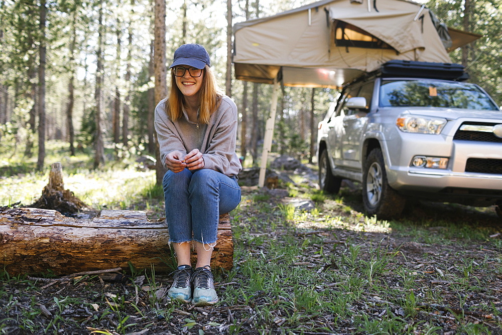 Portrait of smiling woman sitting on log on camping, car with tent in background, Wasatch Cache National Forest