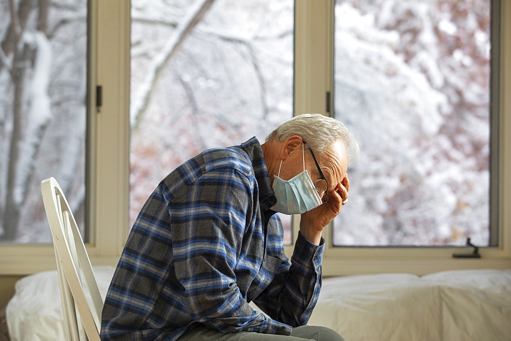 Senior man in Covid protective mask sitting on chair by window