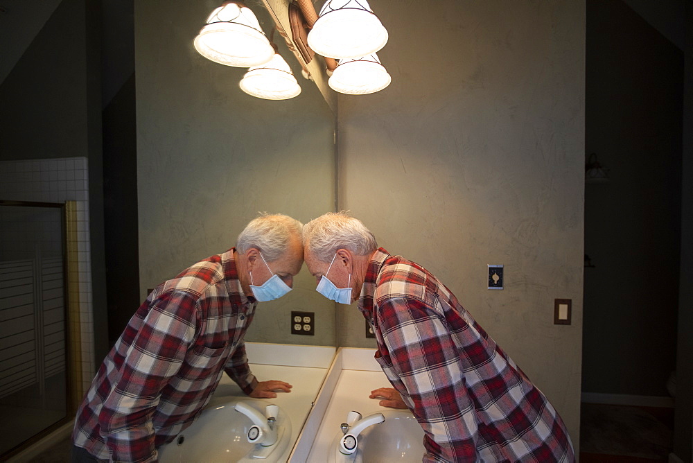 Senior man in Covid protective mask leaning head on mirror in bathroom