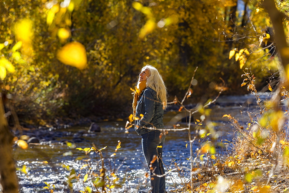 United States, Idaho, Sun Valley, Senior woman standing in sunlight by river in autumn