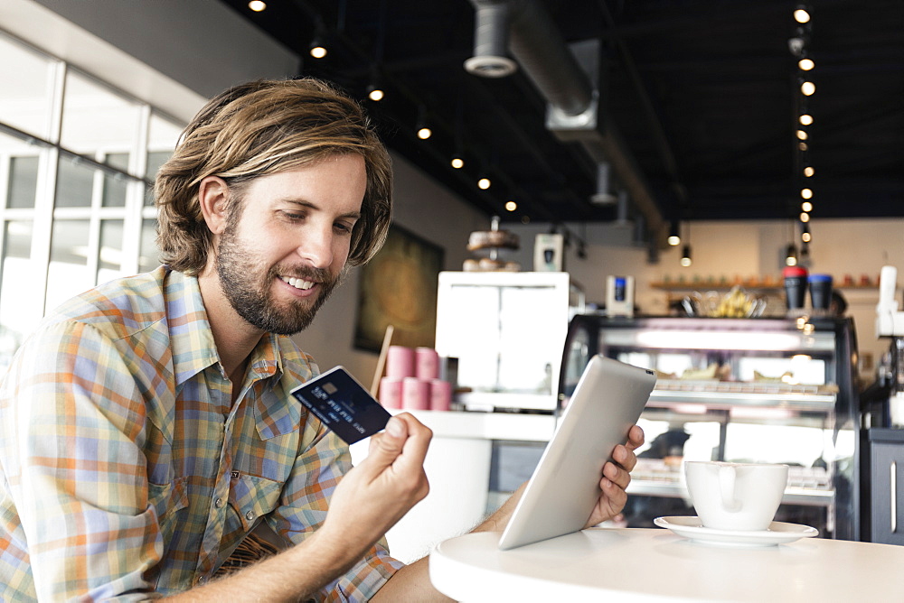 Mid adult man in coffee shop, using digital tablet, holding credit card