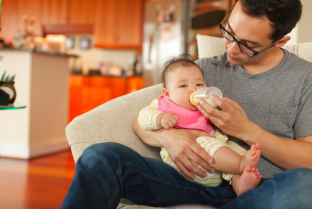 Father sitting on sofa feeding baby daughter