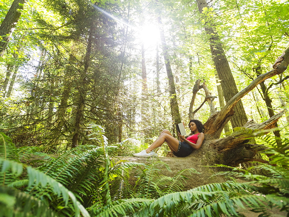 Young woman reading book on log in forest, USA, Oregon, Portland