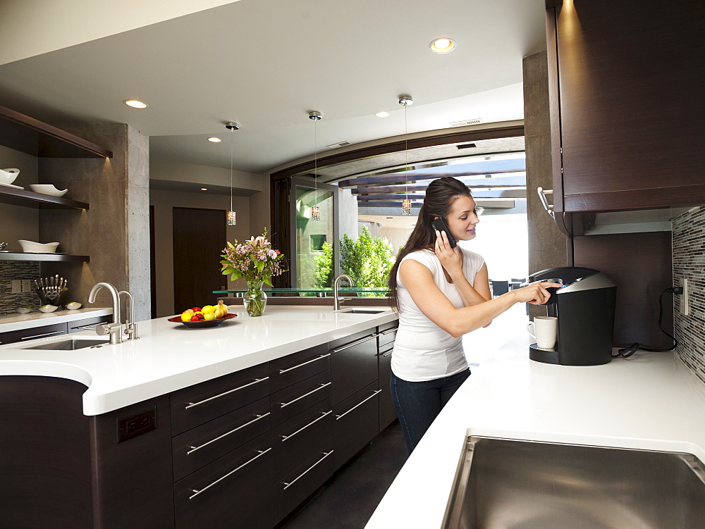 Young woman making morning coffee while using mobile phone