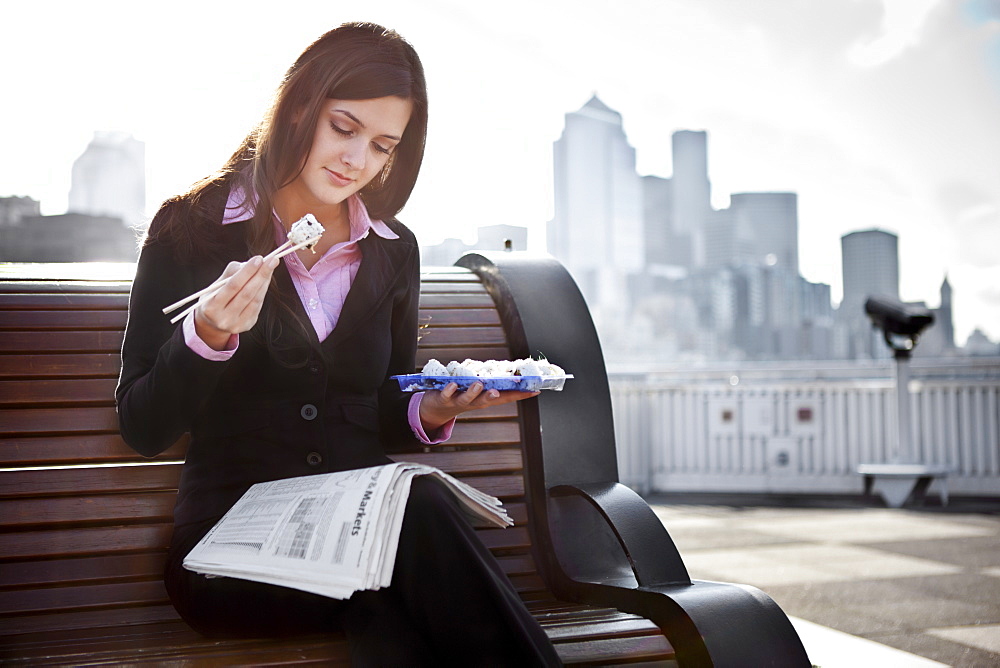 USA, Seattle, Young businesswoman eating sushi and reading newspaper