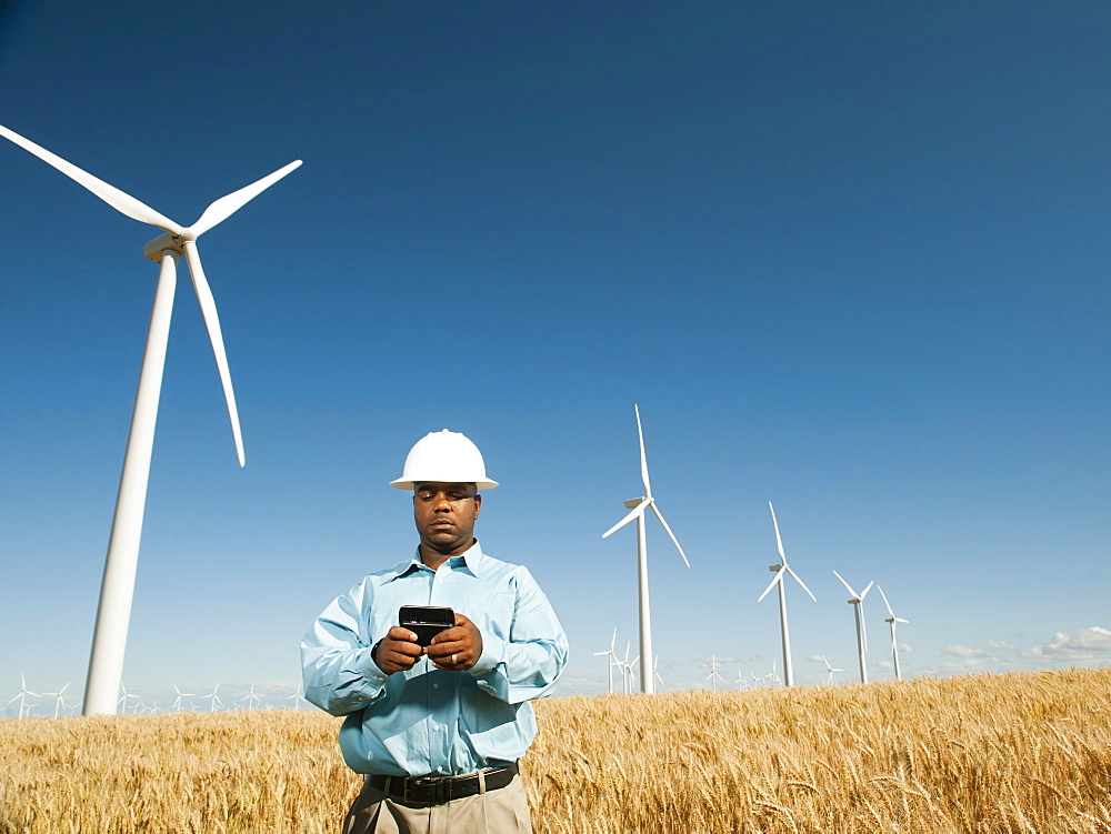 USA, Oregon, Wasco, Engineer standing in wheat field in front of wind turbines, using mobile phone