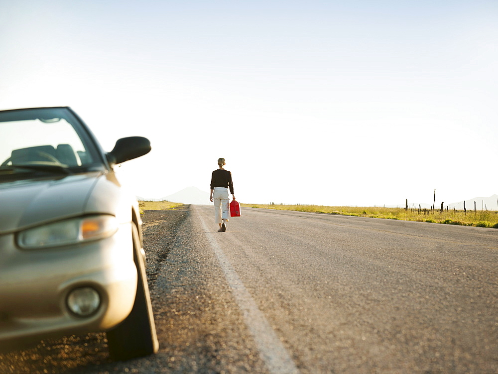 Woman carrying canister walking along empty road, her car parked on roadside