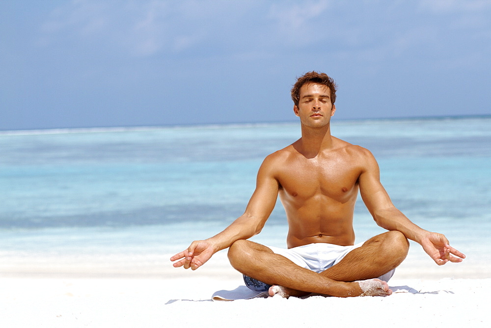 Handsome young man meditating in a lotus position on the beach