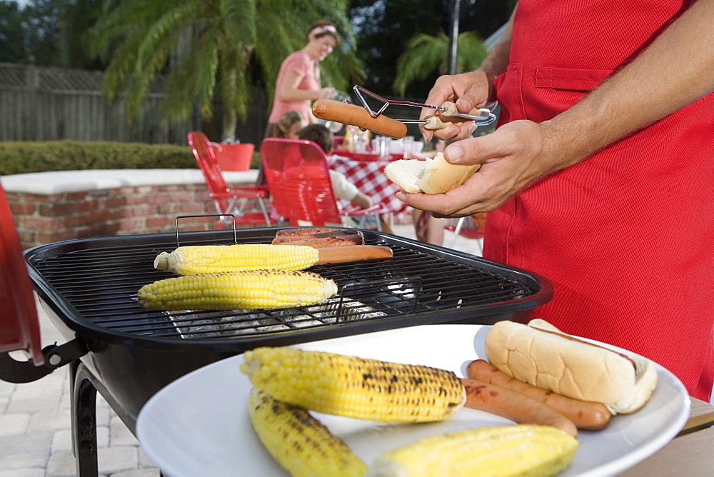 Man grilling hotdogs and corn in backyard