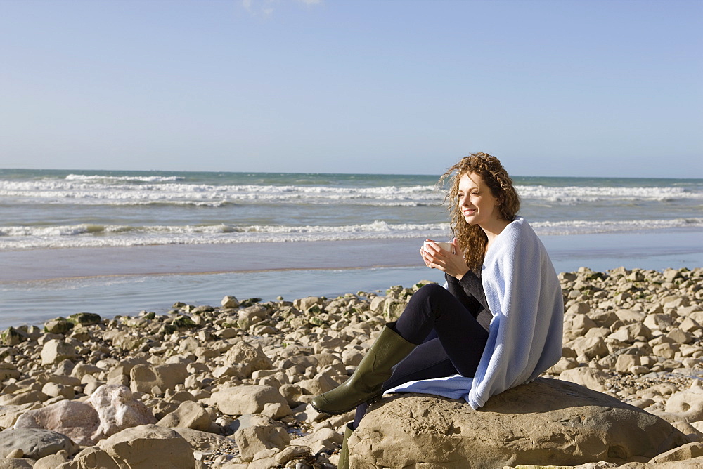 France, Pas-de-Calais, Escalles, Woman wrapped in blanket sitting on rocky beach, France, Pas-de-Calais, Escalles