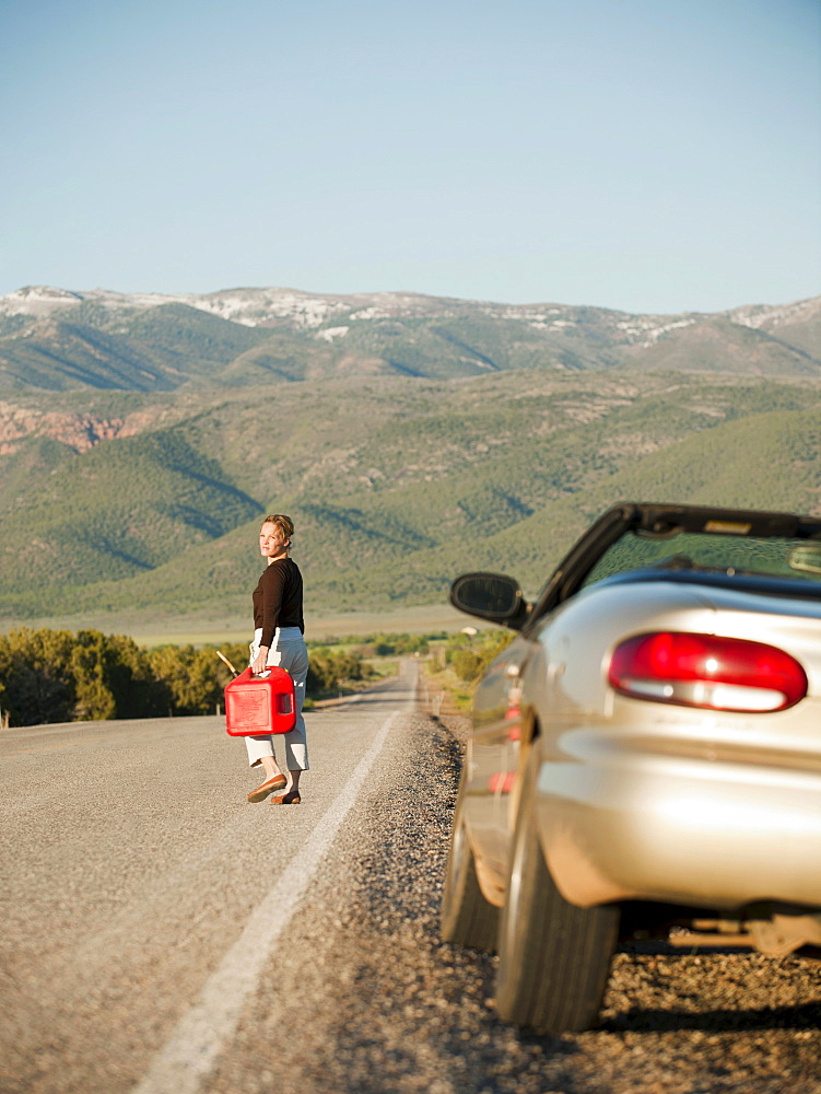 USA, Utah, Kanosh, Woman carrying canister walking along empty road, her car parked on roadside