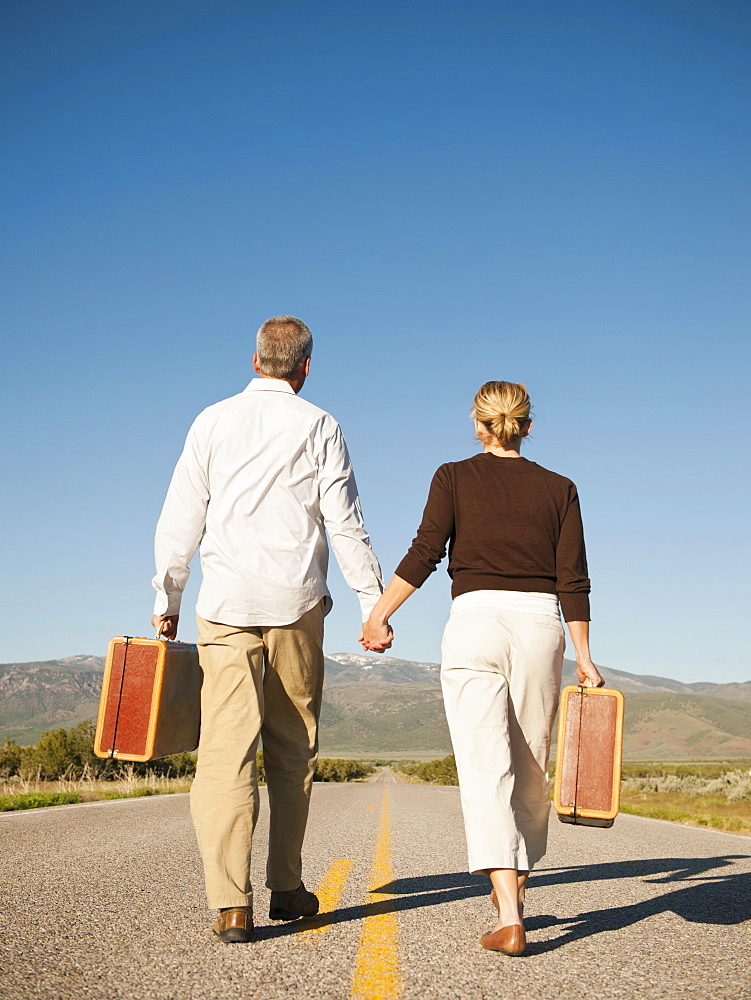 Mid adult couple walking along empty road carrying suitcases