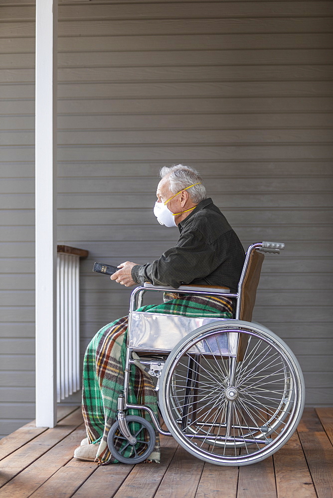 Senior man in wheelchair wearing protective mask to prevent coronavirus transmission on porch