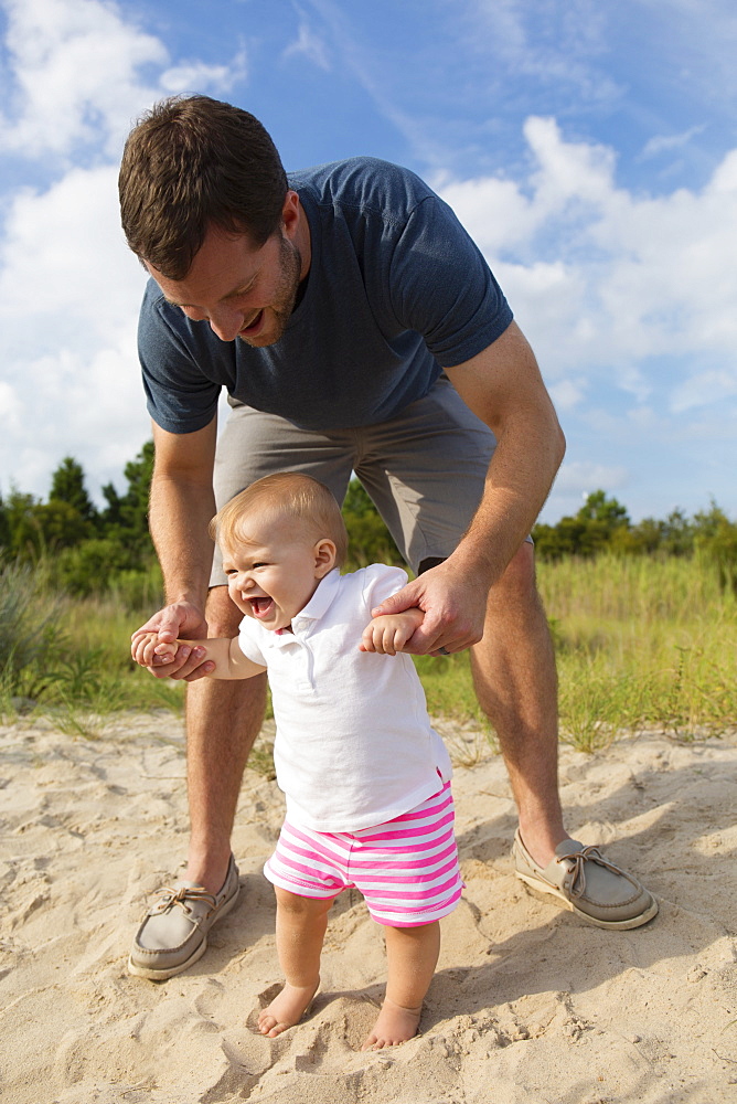 Mid adult man holding baby daughters hands while toddling in sand