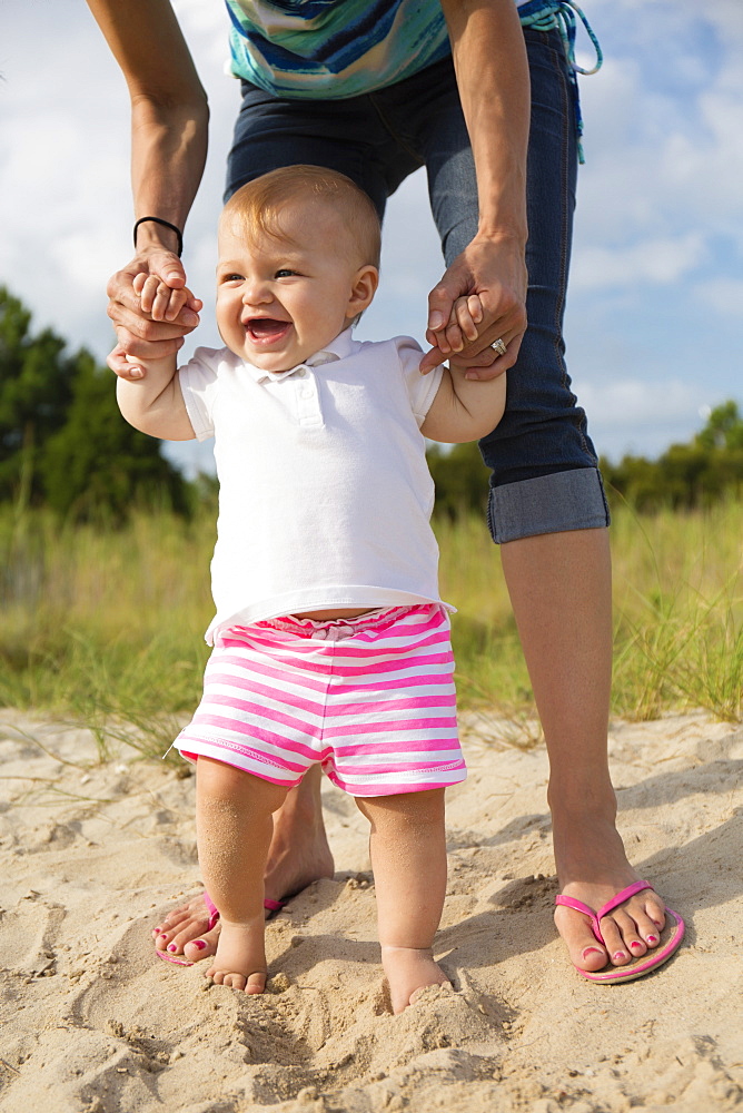 Mid adult woman holding baby daughters hands while toddling in sand