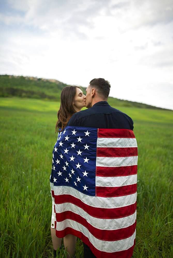 Young couple wrapped in American flag kissing in wheat field