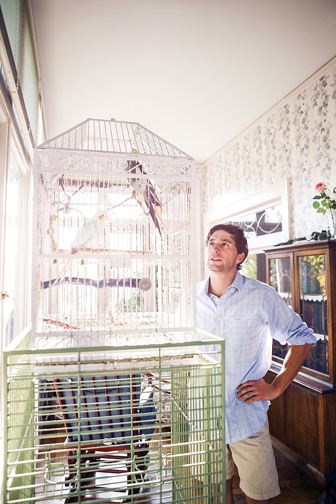 Portrait of young man standing by bird cage and looking at parrot