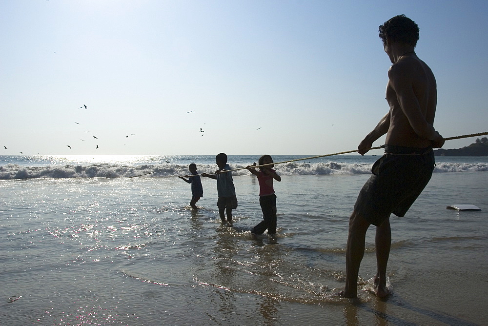 Man and three children hauling in the fishing net on the beach, Goa, India