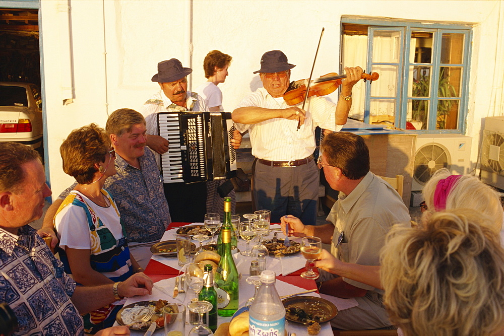 Tourists in open air restaurant being entertained by musicians including man playing accordion on Mykonos, Cyclades Islands, Greek Islands, Greece, Europe