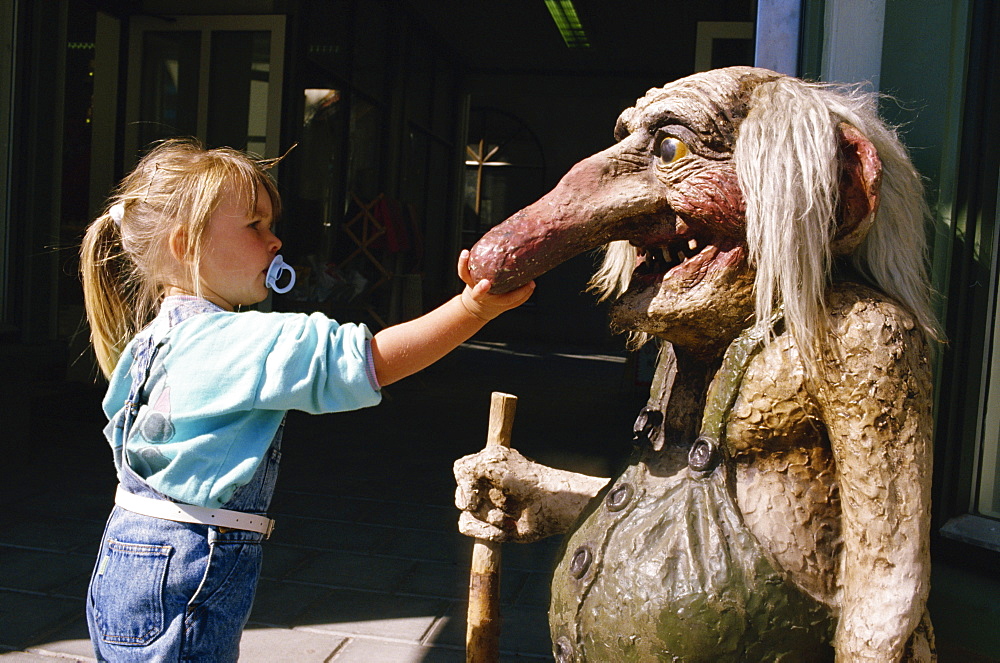 Young girl with dummy in her mouth, touching troll statue with long nose, Norway, Scandinavia, Europe