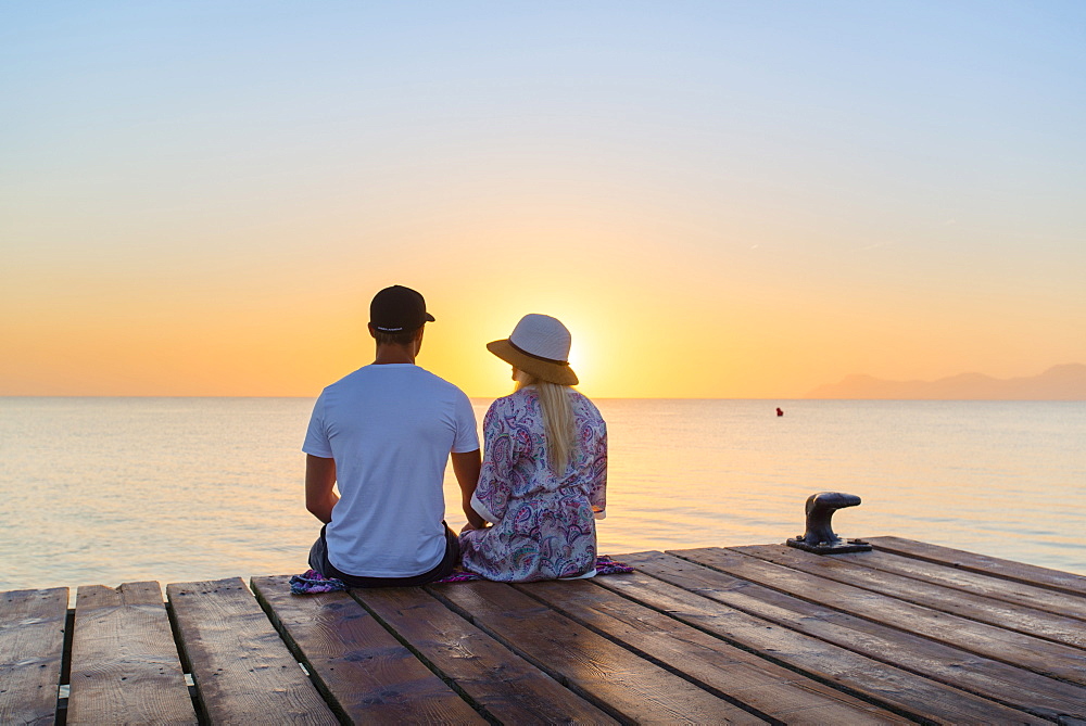 Young couple sitting at the end of a jetty in the morning atmosphere and enjoying the view of the sunrise. Playa de Muro beach, Alcudia, Mallorca, Balearic Islands, Spain