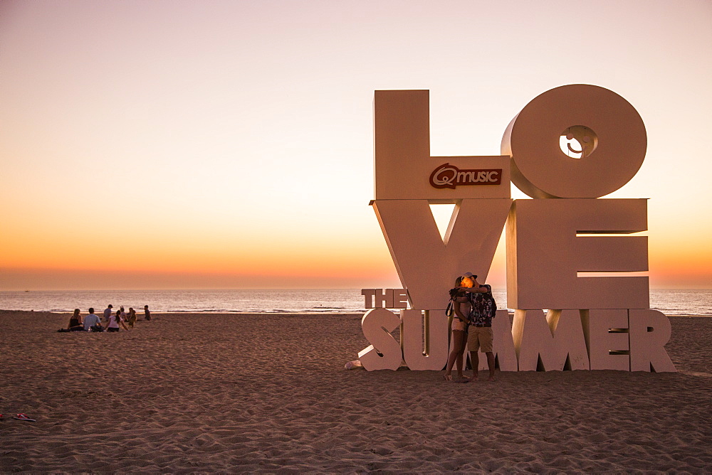 Couple taking a selfie photograph with smartphone in front of Love sculpture on the beach at sunset, Ostend, Flanders, Flemish Region, Belgium