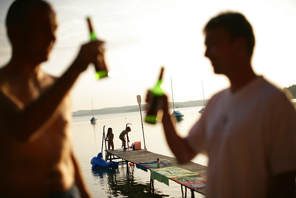 Two men drinking bottles of beer, children playing on jetty in background, Lake Woerthsee, Bavaria, Germany, MR