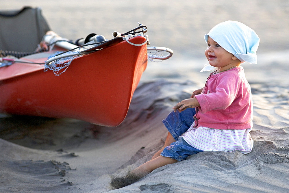 A one year old girl sitting on the beach next to a sea kayak, Punta Conejo, Baja California Sur, Mexico