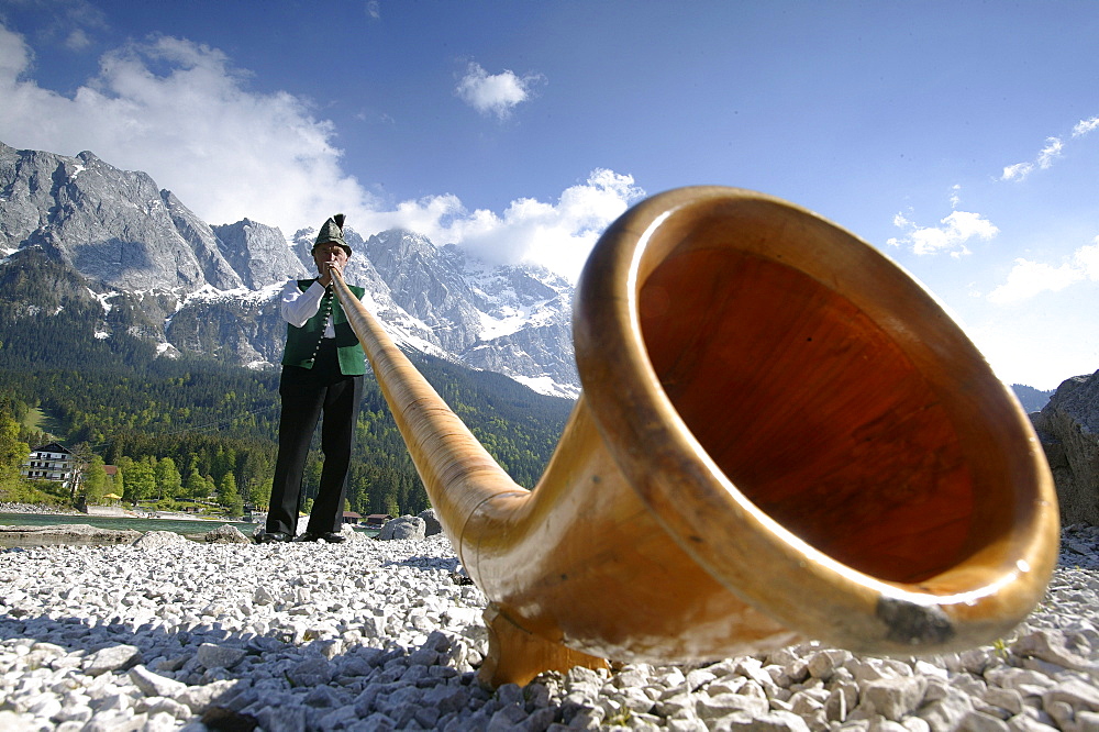 Alphorn blower standing at shore of Lake Eibsee, mount Zugspitze in the background, Garmisch-Partenkirchen, Bavaria, Germany