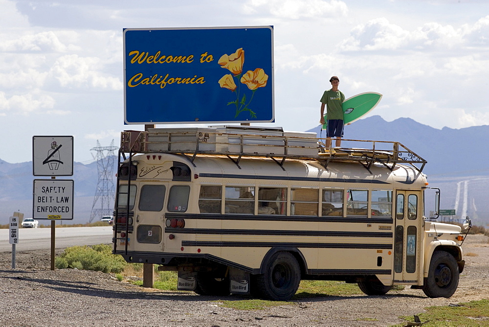 An 18 year old teenager with a surfboard standing on top of an American Schoolbus at the California welcome sign, Interstate 15, Nevada, California, USA