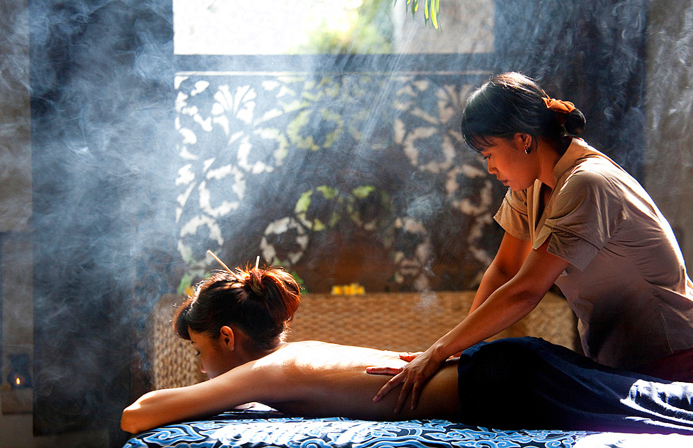 A Balinese therapist performing a massage on an Asian woman, in a smoky, romantic setting. Bali, Indonesia.