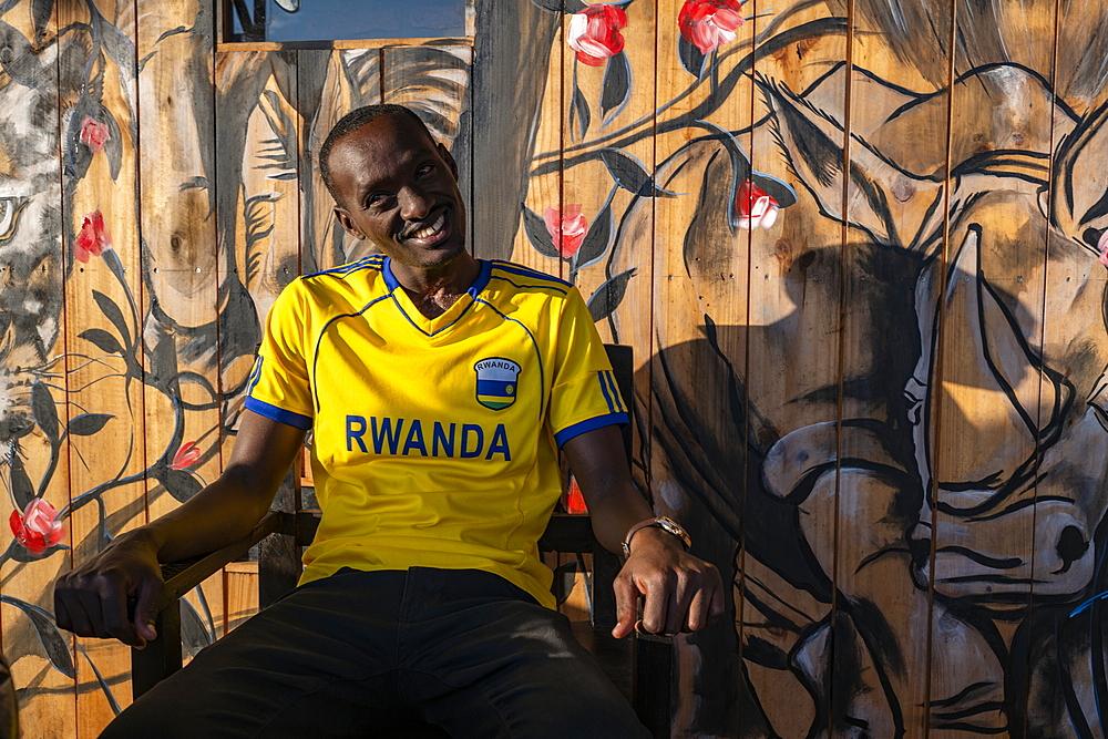 Young man with Rwanda jersey sits in front of mural with safari animal motif in a cafe, Kayonza, Eastern Province, Rwanda, Africa