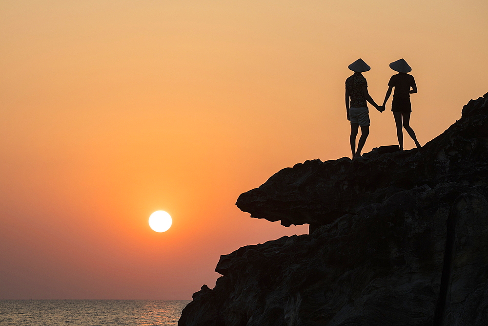 Silhouette of young couple wearing conical hats and looking out to sea from rock ledge next to Dinh Cao Shrine at sunset, Duong Dong, Phu Quoc Island, Kien Giang, Vietnam, Asia