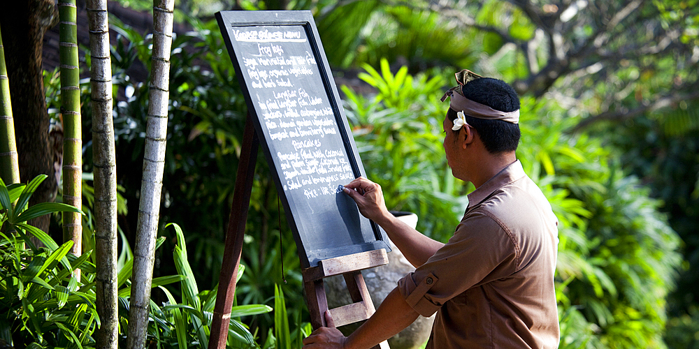 A Balinese waiter writing the daily menu on a blackboard in a tropical setting. Singaraja, Bali.