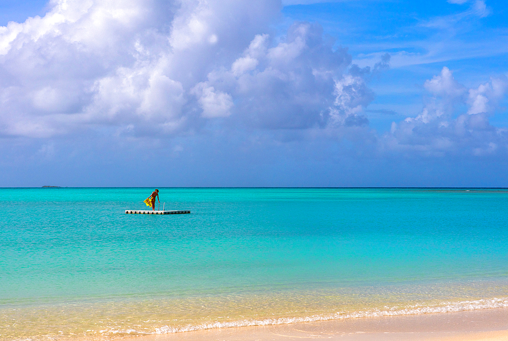 A woman with a bright yellow sarong, standing on a float, off a beach, agains a turqoise sea. Shot in Antigua.