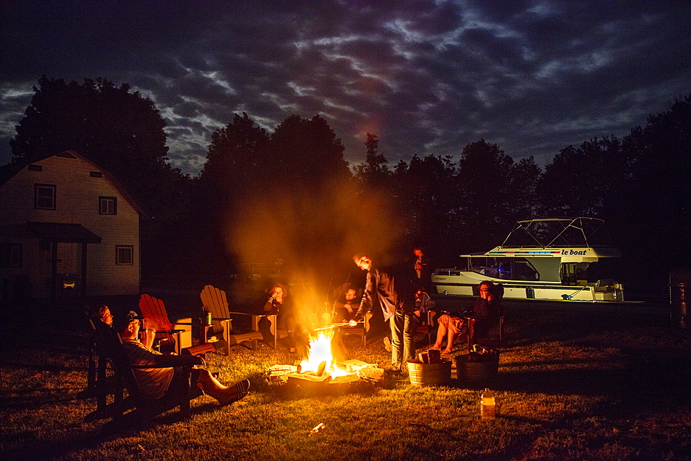 People relax by the campfire under the full moon at Parks Canada Campground at Beveridge Locks on the Tay River with a docked Le Boat Horizon houseboat at night, near Lower Rideau Lake, Ontario, Canada, North America