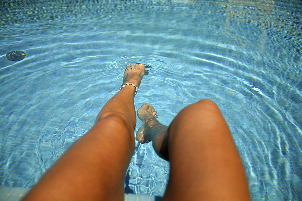 Woman dangling her legs into water, sitting on the edge of a swimming pool