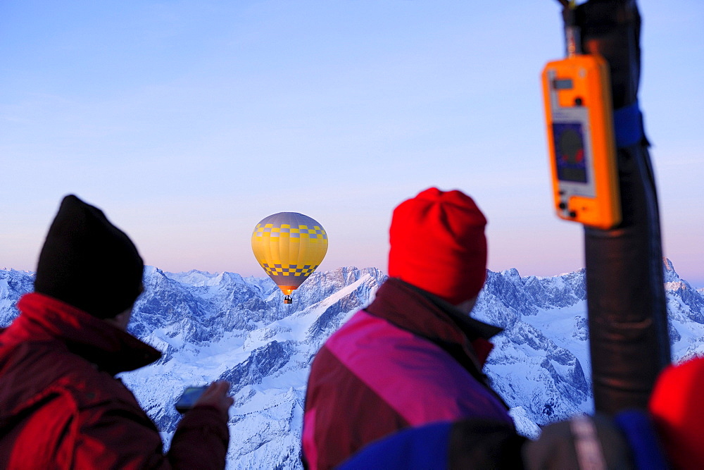 Two people on hot-air ballon ride looking towards hot-air balloon, snow covered mountains in background, Garmisch-Partenkirchen, Wetterstein range, Bavarian alps, Upper Bavaria, Bavaria, Germany, Europe